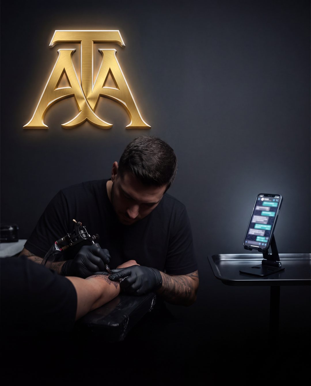 A tattoo artist at work under the glowing TAA monogram, with a phone on the tray showing the WhatsApp chat